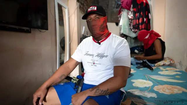 A man with a pistol sits on a bed. He's wearing blue shorts, a white Tommy Hilfiger t-shirt, a black cap and a red balaclava obscuring his face. 