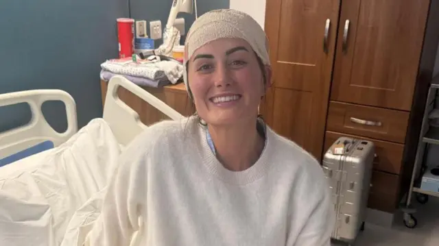 Lauren Macpherson está sentada em uma cama de hospital. Ela tem uma faixa branca enrolada na cabeça, olhos castanhos e cabelo castanho