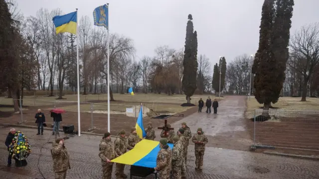 Egyenruhás katonák egy ukrán kék‑sárga zászlót tartanak a koporsó fölé egy gyakorlótéren.