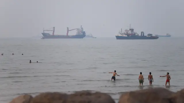 Puslu bir günde, ön planda açık denizde kargo gemileri ve tankerler dururken, insanlar kıyı sularında yüzüyor ve suya giriyor.