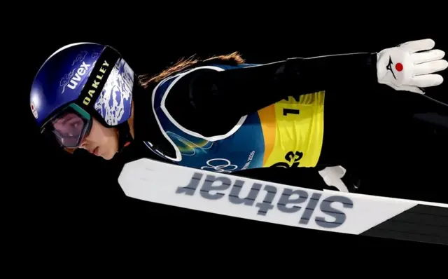 黒いウェアの高梨が空中で深い前傾姿勢を取りながら前方を見ている