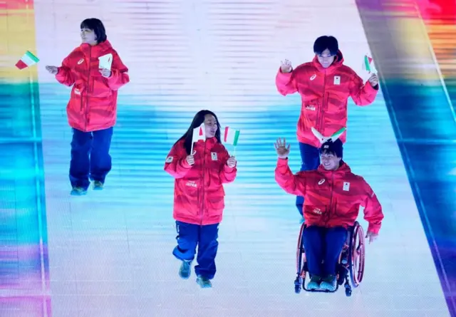 上半身は赤色、下半身は紺色のそろいのウエアを着て日本とイタリアの小旗をもった4人が照明で白く照らされた通路を行進している。1人は車いすに乗っている