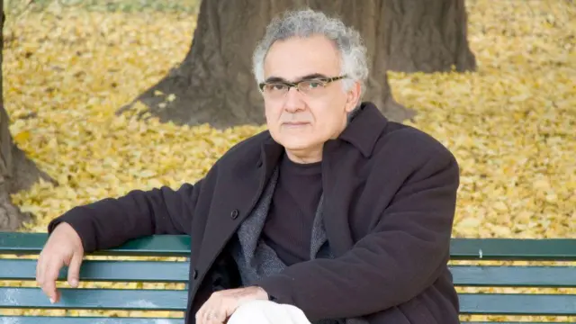 Milton Hatoum, um homem de cabelos grisalhos e óculos, aparece sentado em um banco de parque, com expressão séria e contemplativa. Ao fundo, o chão está coberto por folhas amarelas. Aparecem ainda os troncos das árvores.