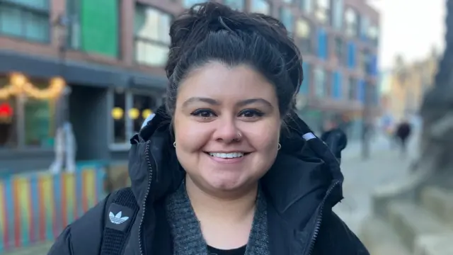 Una mujer con el pelo negro recogido en lo alto de la cabeza sonríe. Lleva un abrigo negro con capucha.