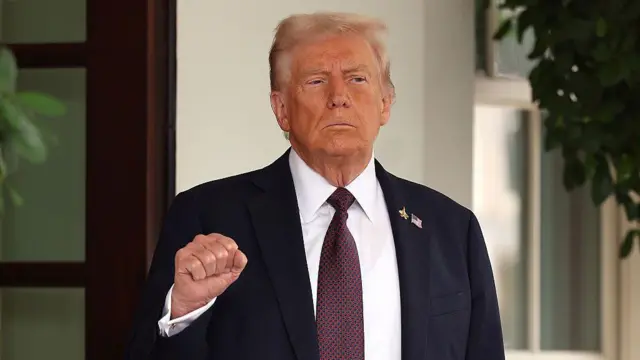 Donald Trump con una chaqueta oscura, camisa blanca y corbata vino tinto. Levanta el puño derecho cerrado a la altura del pecho.