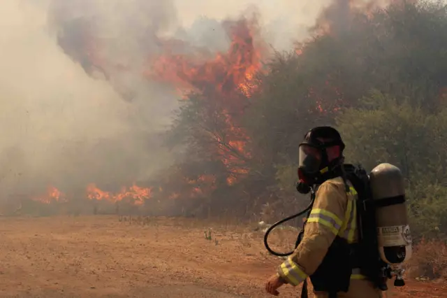 Flames a smoke billow in the air as a suited and masked firefighter prepares to put the blaze out