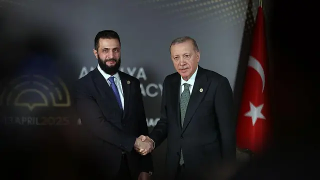 Cumhurbaşkanı Recep Tayyip Erdoğan ve Suriye Cumhurbaşkanı Ahmed Şara iki ülke bayrakları arasında el sıkışıyor.