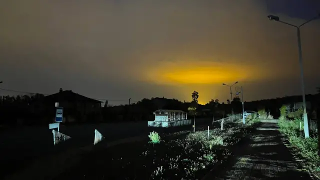 В темноте можно разглядеть знак автобусной остановки и саму остановку, а также дорогу, ведущую в направлении леса