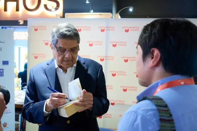 Una foto del escritor Sergio Ramírez autografiando un libro. Frente a él está un hombre que se ve de espaldas a la cámara.