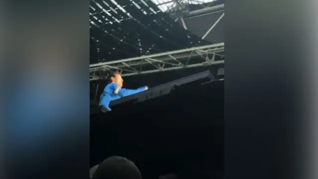 Zrzut ekranu z występu Harry’ego Stylesa na scenie, gdzie wysokie elementy scenografii częściowo zasłaniają widok na artystę ubranego na niebiesko.