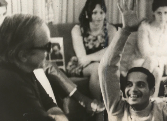 Foto em preto e branco de 1972 mostrando Baden Powell e Vinicius de Moraes em momento descontraído. Vinicius está sentado, de óculos escuros, sorrindo, enquanto Baden Powell, à frente, levanta o braço em gesto animado. Ao fundo, algumas pessoas observam a cena em um ambiente informal.