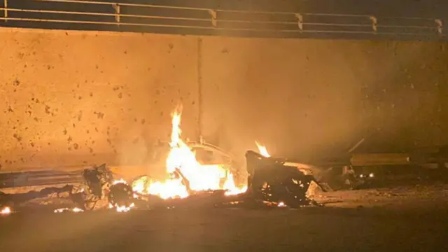 A handout photo made available by Iraq's military-run Security Media Cell via Facebook shows the remains of a burning vehicle beside a tall concrete wall. The hulk of wheels and the bonnet can be made out, with the main bulk of the vehicle in flames. The tall wall with railings on the top is behind the vehicle. It is night and orange flames illuminate the area.