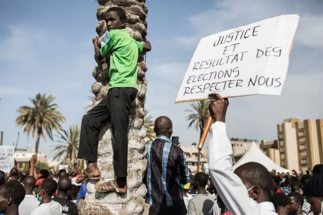 Un jeune homme en t-shirt vert et pantalon noir grimpe le tronc d'un arbre, un homme tient à côté de lui une pancarte sur laquelle est écrite :"Justice et résultat des élections, respectez-nous".une foule de personnes est en arrière-plan.