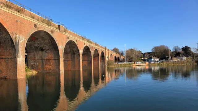 Un día soleado con un cielo azul brillante muestra un viaducto que discurre por el lado izquierdo de la imagen. Un cuerpo de agua corre junto al viaducto, lo que se refleja en la superficie.