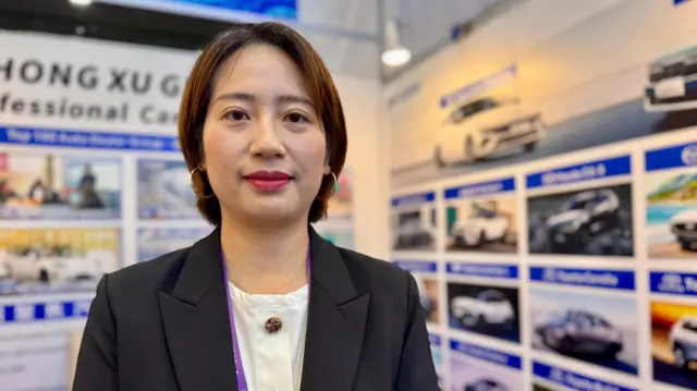 Joyce Liu bir elektrikli araç standında. Kısa kahverengi saçlı Joyce, beyaz bir gömlek ve şık siyah bir ceket giymiş, doğrudan kameraya bakıyor.