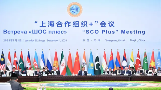 天津上海合作组织峰会习近平坐在会议厅巨型圆桌中央，背后是与会各国国旗旗海和上合组织标志布景板（1/9/2025）