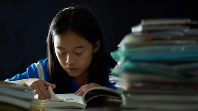 Menina asiática lendo um livro, com uma pilhabetpix365 tigrecadernos sobre a mesa