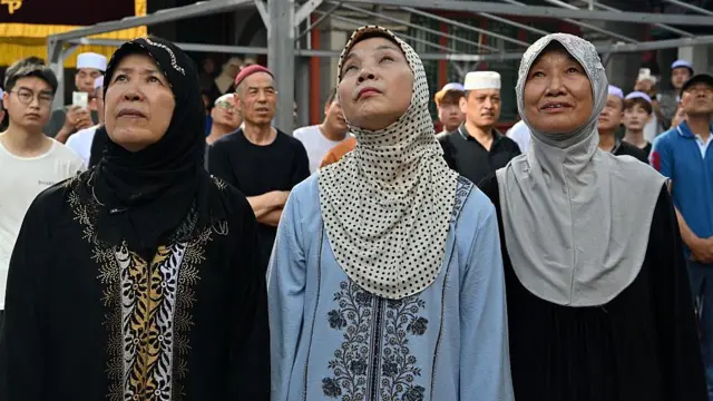 Three women wit hijab looking up. Some men also dey di background.