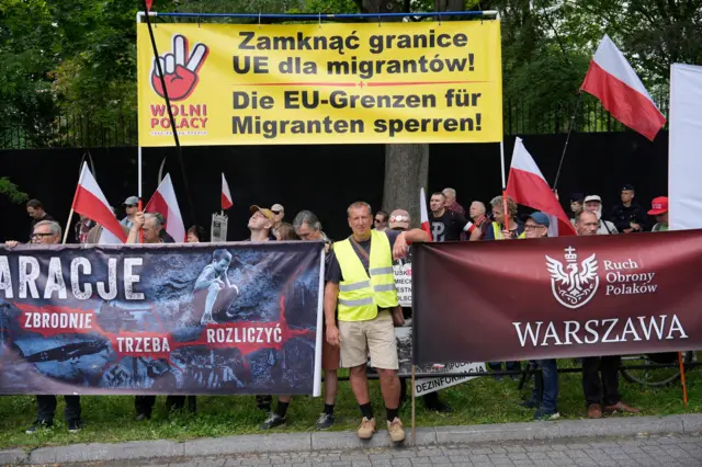 Grupa Polaków pikietująca na granicy z Niemcami przeciwko zawracaniu migrantów do Polski. Protestujący trzymają biało-czerwone flagi i trzy banery, jeden z nich w kolorze żółytym, z hasłem wzywającym do zamknięcia granicy. Pomiędzy dwoma banerami stoi mężczyzna w kamizelce odblaskowej, Porajów, 3 lipca 2025.