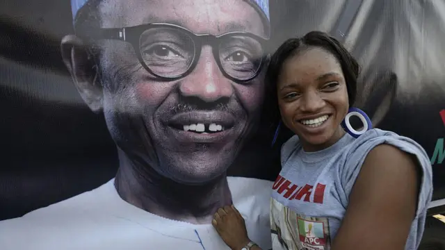 Une femme pose à côté d'une affiche représentant un Buhari souriant. Elle porte un t-shirt gris sur lequel sont inscrits « APC » et « Buhari ».