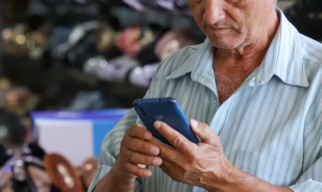 Idoso usando um celular. Ele veste camisa listrada azul clara e o celular tem capinha azul, seu rosto é visto apenas da metade para baixo