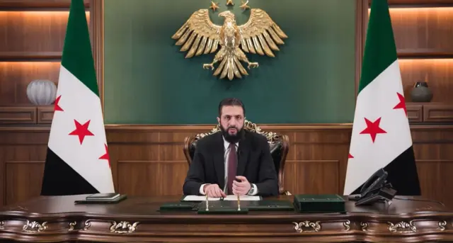 Suriye lideri Şara, Suriye bayraklarının önünde kurulmuş bir masada açıklama yaparken. 