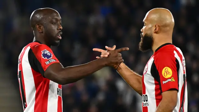 Yoane Wissa et Bryan Mbeumo, tous deux vêtus du maillot rouge et blanc de Brentford, se font face et se serrent la main, tout en se désignant du doigt avec l'index de la même main, lors d'un match disputé sous les projecteurs.