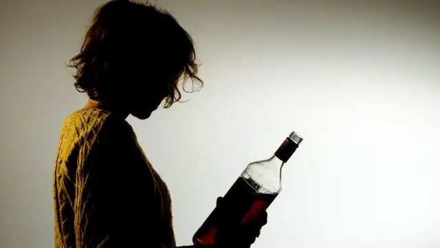 Slika devojke u žutom džemperu iz profila sa flašom alkohola u ruci 