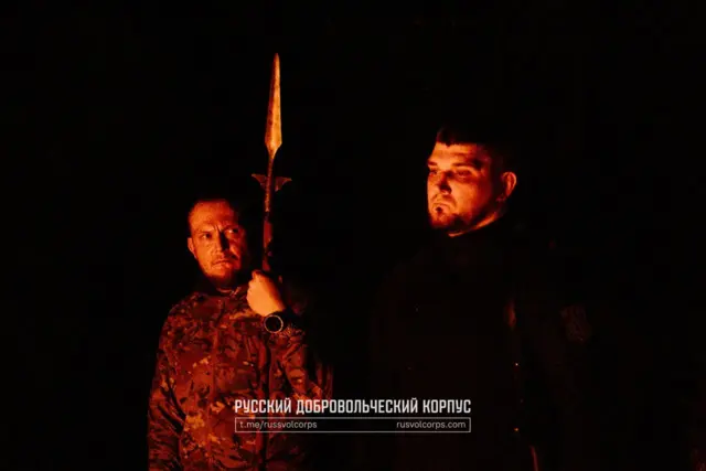 Двоє військових, освітлені полум’ям у темряві: один стоїть ліворуч у камуфляжі й тримає довгий спис або обрядову спицю вертикально перед собою. Інший – праворуч, у темному одязі, з нашивкою на рукаві. Обличчя обох освітлює червоно‑помаранчеве світло вогню, що створює драматичний, похідний настрій.
