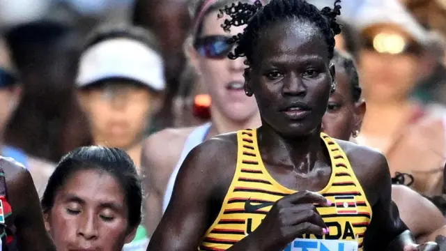 Ugandalıkadın atlet Rebecca Cheptegei, Budapeşte'de "UGANDA" yazılı sarı çizgili bir tişört giyerek yarışıyor; arka planda birçok koşucunun yüzleri bulanıklaştırılmış şekilde görülebiliyor.
