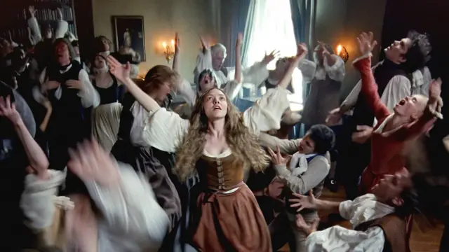 Жінка у старовинному одязі в центрі кадру розводить руки в танці, довкола неї так само емоційно жестикулюють багато людей