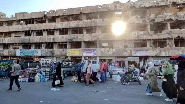 Humus kent merkezinde yıkılmış bir binanın önünde kurulan pazar ve insanlar var