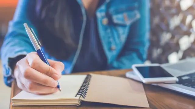 Mulher sentada escrevendo em caderno ao lado de celular e laptop na mesa