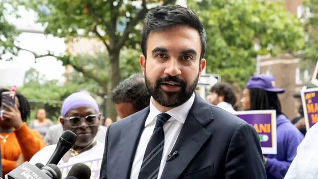 زهران ممداني يتحدث خلال إحدى فعاليات حملته الانتخابية وقد وقف وراءه أعضاء بنقابة العاملين في القطاع الصحي يرفع بعضهم لافتات مؤيده له