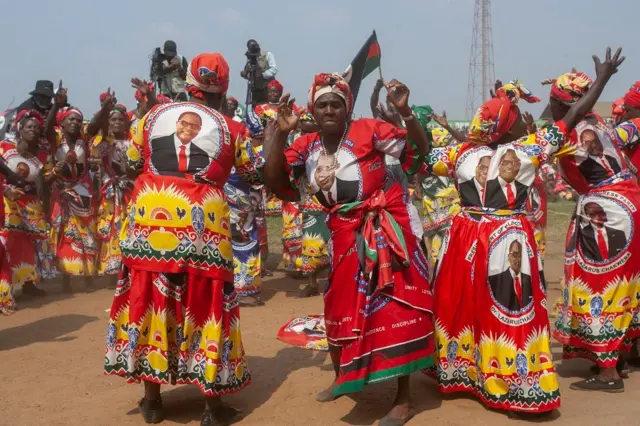 Women wearing matching red dresses bearing the president's face dance in circles.