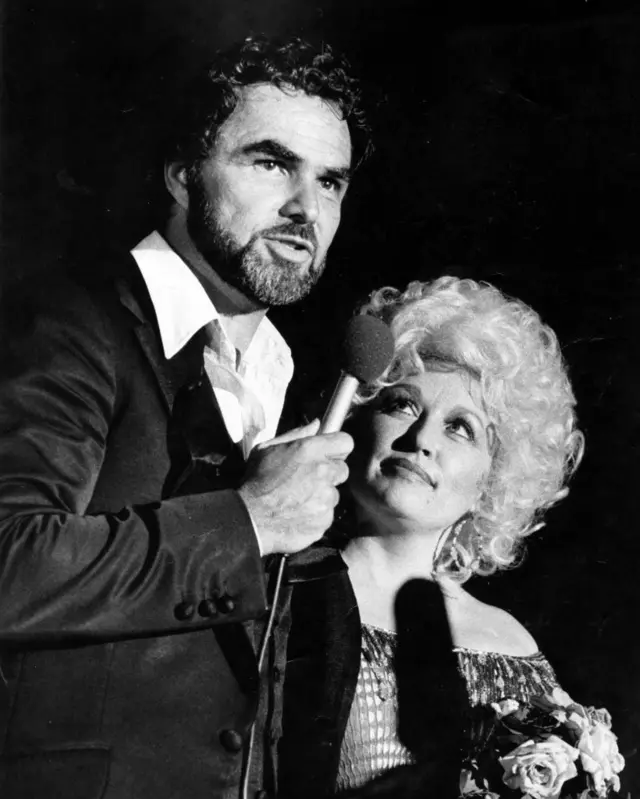 Reynolds abrazado con Dolly tiene un micrófono en mano, ella lo mira con admiración.