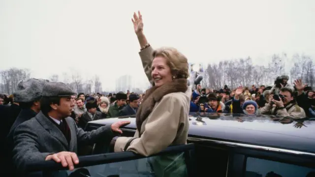 Маргарет Тэтчер машет рукой высунувшись из открытой двери машины приветствуя советских людей пришедших увидеть ее. Двое охранников заслоняют ее спинами от толпы.
