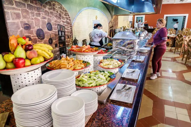 Uma mulher em uma estação de buffet de café da manhã. Há várias frutas e alimentos