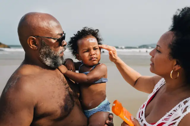 Criança no colotipminer arbetyhomem, enquanto mulher passa protetor solar no rosto da menina