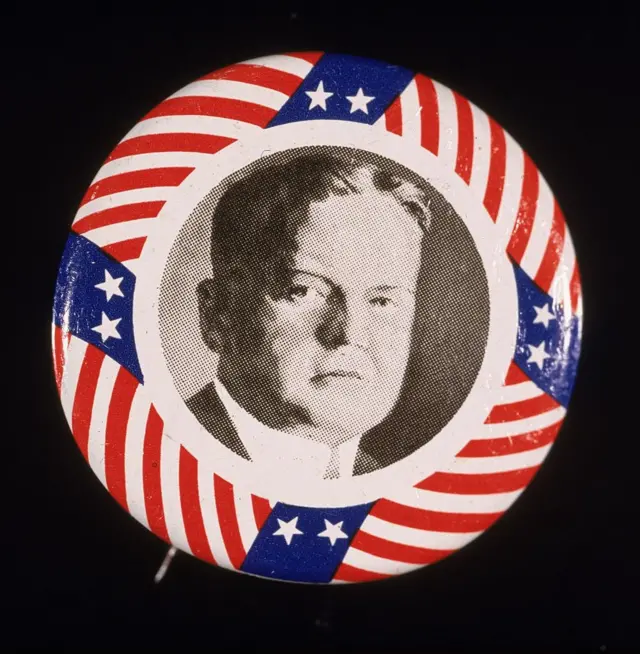 Botón de campaña presidencial con el rostro de Hoover en blanco y negro en el centro y rayas rojas y blancas, así como franjas azules con dos estrellas blancas, rodeándolo.