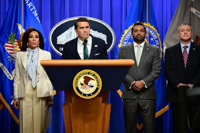 Тодд Бланш у темному костюмі стоїть перед трибуною. Поруч з ним стоїть жінка в білому і чоловіки в сірому і темному костюмах 