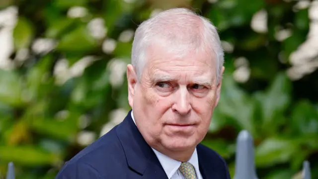 Prince Andrew, a middle aged man with short white hair, is dressed in a navy suit with a white shirt and yellow and green tie.