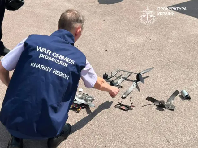 На передньому плані людина у жилеті з написом "WAR CRIMES prosecutor KHARKIV REGION" розглядає розкладені на асфальті фрагменти дрона, серед яких видно елементи корпусу, пропелери та електронні компоненти. Фото документує огляд речових доказів після атаки безпілотника.