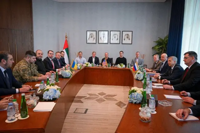 За довгими столами навпроти одна одної сидять делегації України (ліворуч) і Росії (праворуч). У центрі між ними – делегація США. На стіні за нею висять три портрети арабських лідерів у рамках. Ліворуч від них стоїть великий прапор Об’єднаних Арабських Еміратів на золотистому держаку.