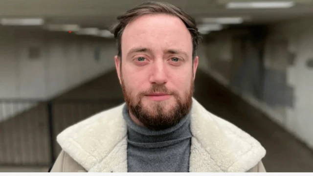 Jacob tem barba e está usando um suéter cina e uma jaqueta bege por cima