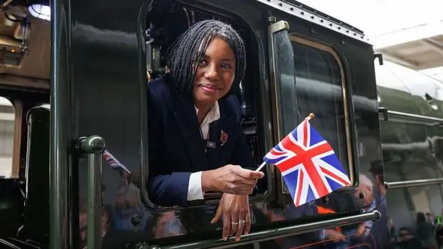 Кеми Баденок выглядывает из окна паровоза, держа в руках британский флажок