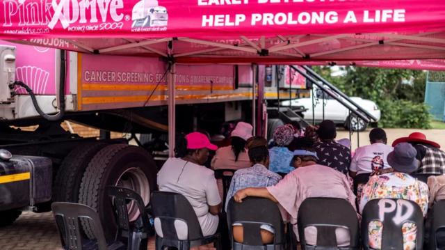 Mulheres sentadas ao lado de van rosa de campanha para realizar exame para detecção precoce de câncer 