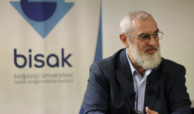 Nureddin Yıldız Boğaziçi Boğaziçi Üniversitesi İslam Araştırmaları Kulübü'nün logosu önünde kürsüde oturuyor. Beyaz uzun bir sakalı ve kravatsız bir takım giyiyor.
