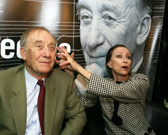 Щедрин в костюме с галстуком сидим и смотрит вдаль, Плисецкая в пиджаке в клеточку держит руки с растопыренными пальцами около его головы.