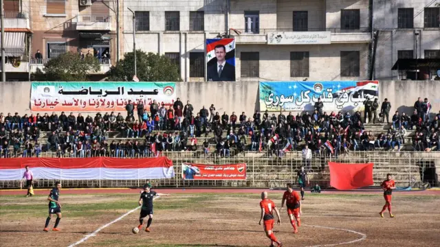 The stand by the sidelines is seen with a crowd - while a banner showing Syrian President Bashar Al-Assad hangs in the background